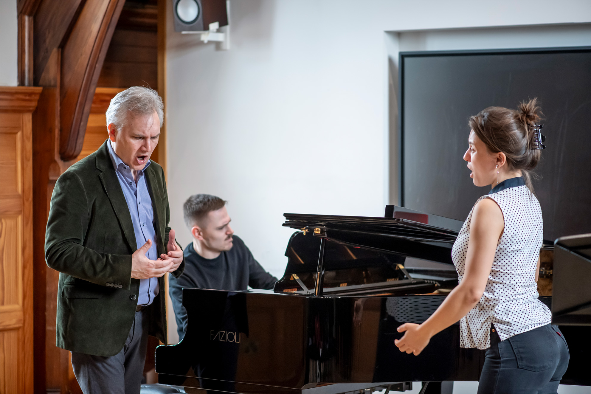 A man guiding and singing alongside a student singing, as a pianist performs on a piano in a wooden panelled room.